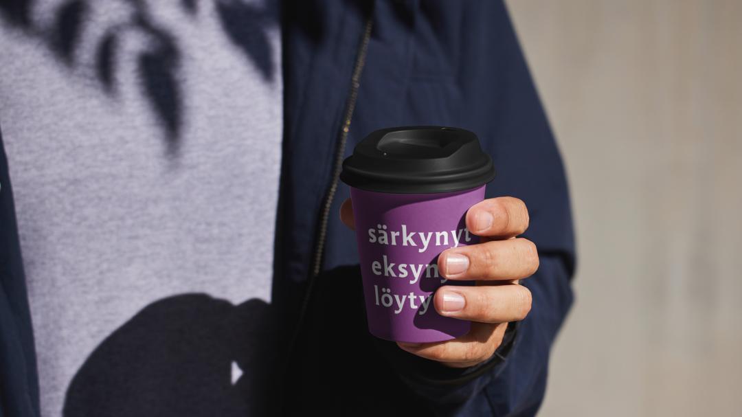 henkilö osittain kuvattuna kädessään lila take away muki