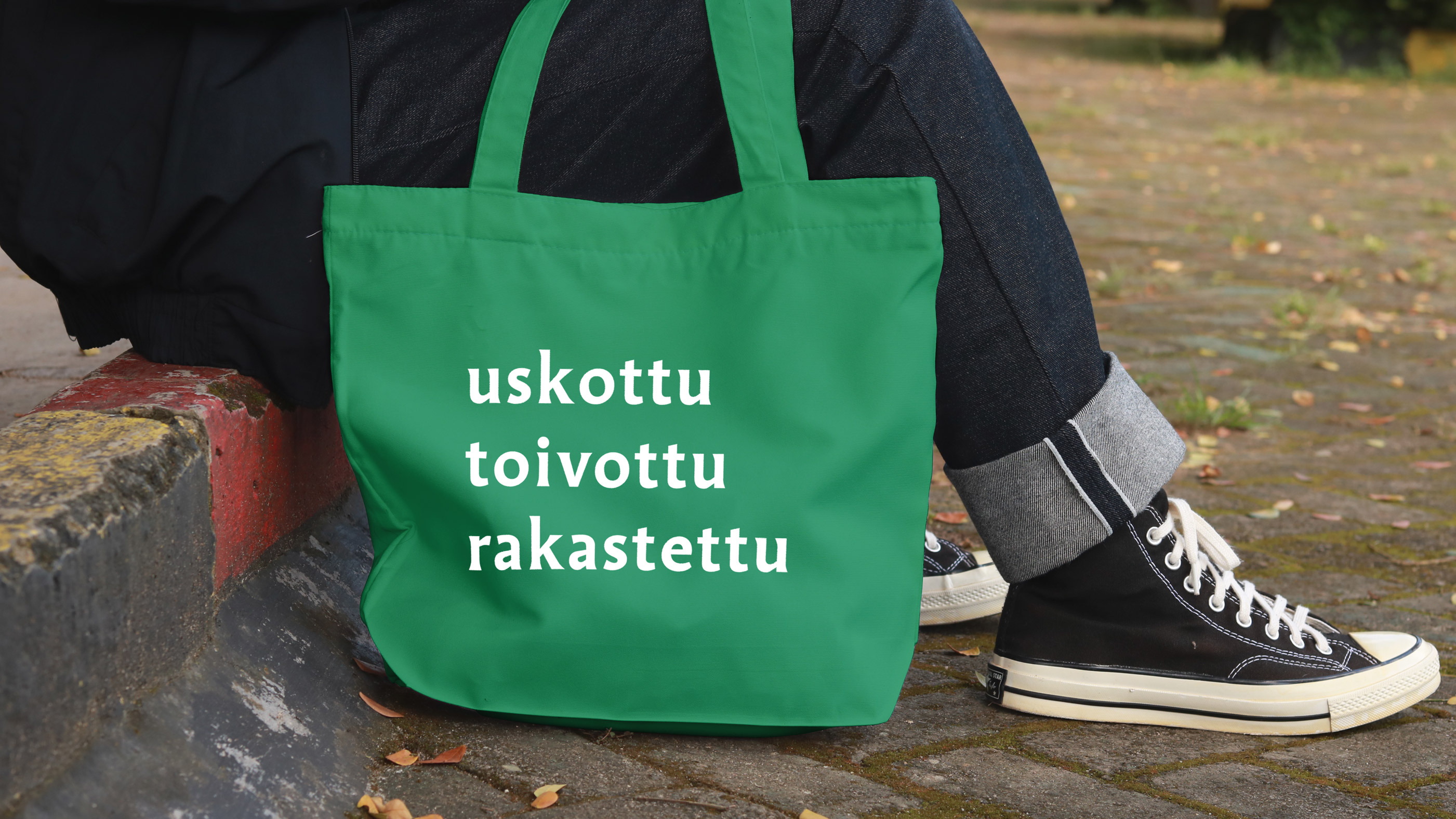 Farkkuihin ja tennareihin pukeutunut henkilö istuu katukivetyksellä vieressään vihreä kangaskassi
